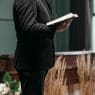 a person wearing a black suit holding a book near a brown coffin